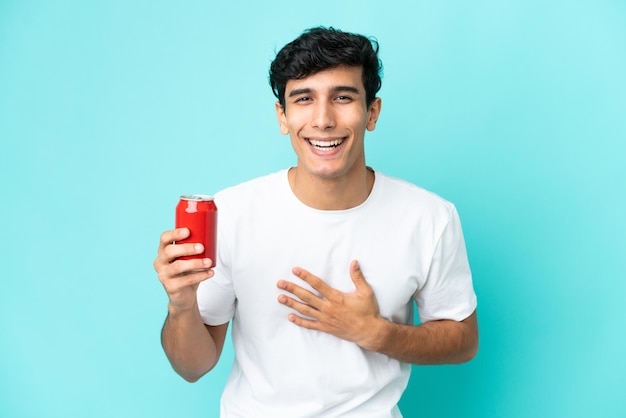 Joven argentino sosteniendo un refresco aislado sobre fondo azul sonriendo mucho