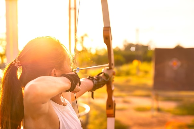 Joven arquera tiro con arco dispara con arco en el campo de la naturaleza para apuntar al concepto de éxito en el campo para el ejercicio deportivo al atardecer