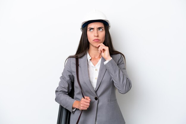 Foto joven arquitecta caucásica con casco y sosteniendo planos aislados en fondo blanco con dudas y pensando