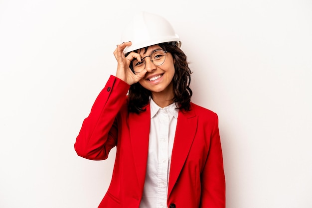 Joven arquitecta hispana con casco aislado en fondo blanco emocionada manteniendo el gesto de ok en el ojo