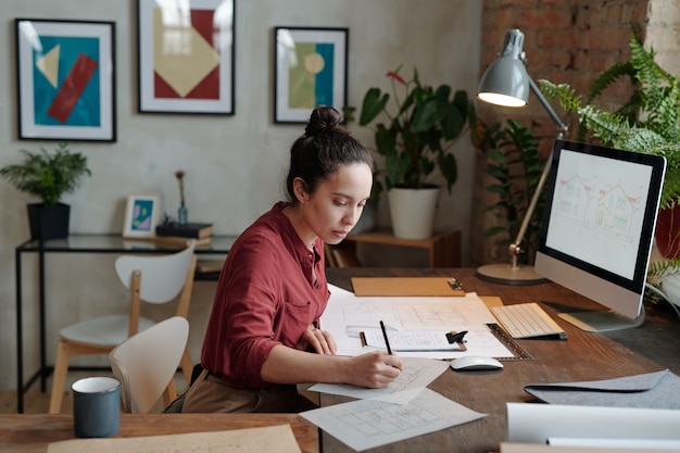 Joven arquitecta seria con dibujo a lápiz boceto sobre papel mientras está sentado junto a la mesa frente a la pantalla de la computadora con la imagen de la casa