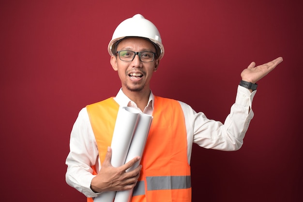 Joven arquitecto asiático con casco y sosteniendo el plan de papel del proyecto apuntando con la mano hacia el lado