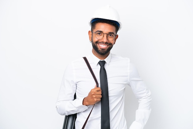 Joven arquitecto brasileño con casco y sosteniendo planos aislados de fondo blanco posando con los brazos en la cadera y sonriendo