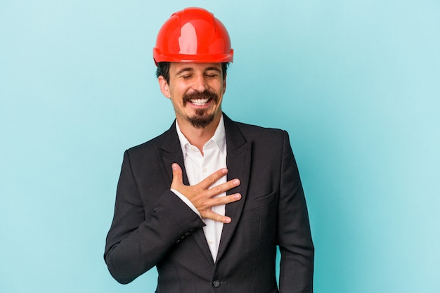 Joven arquitecto caucásico hombre aislado sobre fondo azul se ríe a carcajadas manteniendo la mano en el pecho.