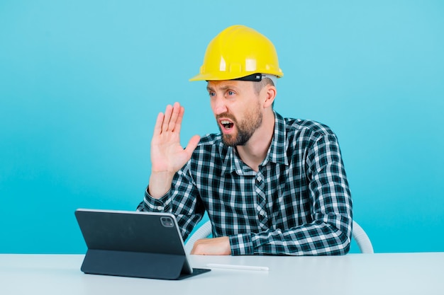 El joven arquitecto grita sosteniendo la mano cerca de la boca con fondo azul.