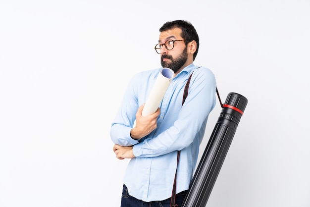 Joven arquitecto hombre con barba sobre pared blanca aislada haciendo dudas gesto mientras levanta los hombros