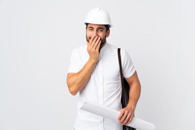 Joven arquitecto hombre con casco y sosteniendo planos aislados en la pared blanca feliz y sonriente cubriendo la boca con las manos