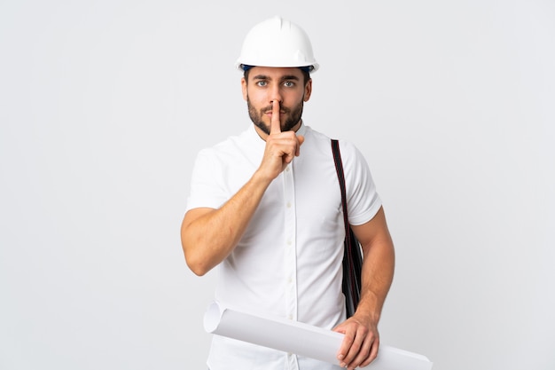 Joven arquitecto hombre con casco y sosteniendo planos aislados en la pared blanca que muestra un gesto de silencio poniendo el dedo en la boca