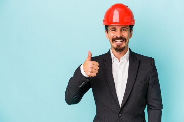 Joven arquitecto hombre caucásico aislado sobre fondo azul sonriendo y levantando el pulgar hacia arriba