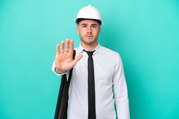 Joven arquitecto hombre caucásico con casco y sosteniendo planos aislados en fondo azul haciendo gesto de parada