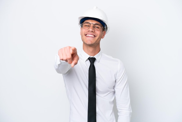 Joven arquitecto hombre caucásico con casco y sosteniendo planos aislados en fondo blanco apuntando al frente con expresión feliz