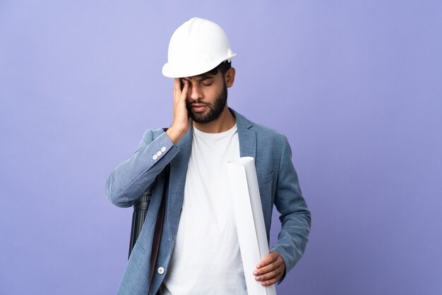 Joven arquitecto marroquí con casco y sosteniendo planos sobre aislados con dolor de cabeza