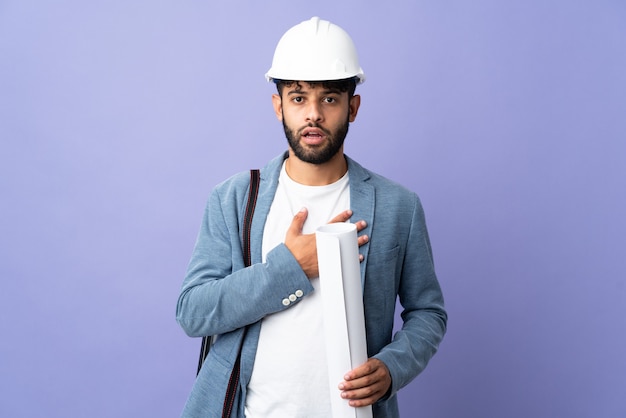 Joven arquitecto marroquí con casco y sosteniendo planos sobre pared aislada sorprendido y conmocionado mientras mira a la derecha