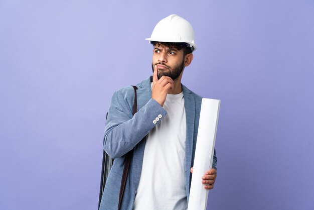 Joven arquitecto marroquí con casco y sosteniendo planos sobre pared aislada teniendo dudas mientras mira hacia arriba