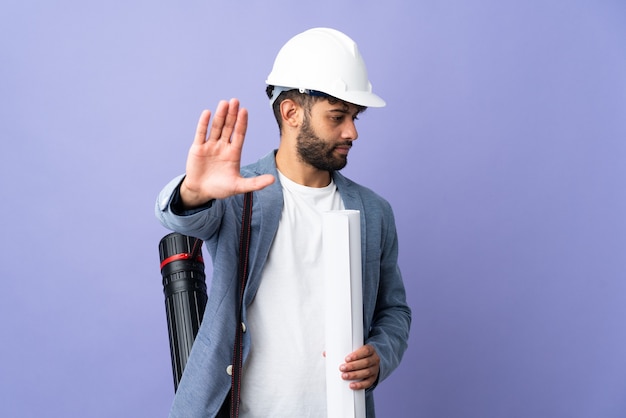 Joven arquitecto marroquí hombre con casco y sosteniendo planos sobre fondo aislado haciendo gesto de parada y decepcionado