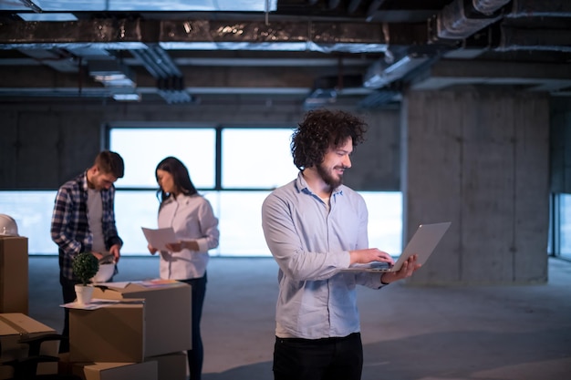 joven arquitecto masculino en el sitio de construcción revisando documentos y flujo de trabajo de negocios usando una computadora portátil con colegas en segundo plano en una nueva oficina de inicio