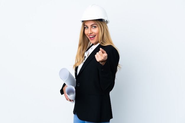 Joven arquitecto mujer con casco y sosteniendo planos sobre la pared blanca señala con el dedo