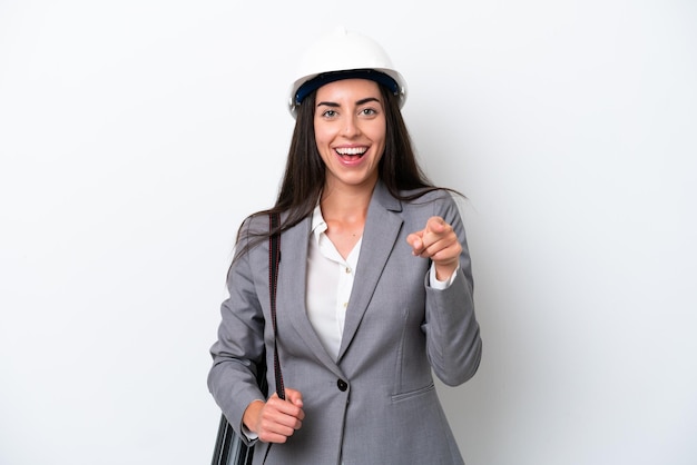 Joven arquitecto mujer caucásica con casco y sosteniendo planos aislados sobre fondo blanco sorprendido y apuntando al frente