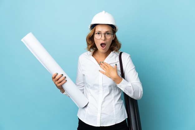 Joven arquitecto mujer georgiana con casco y sosteniendo planos sobre pared aislada sorprendido y conmocionado mientras mira a la derecha