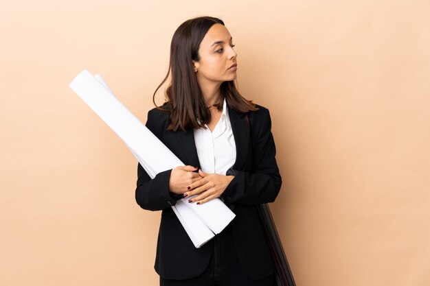 Joven arquitecto mujer sosteniendo planos sobre pared aislada manteniendo los brazos cruzados