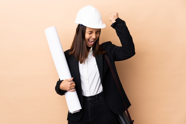 Joven arquitecto mujer sosteniendo planos sobre pared celebrando una victoria