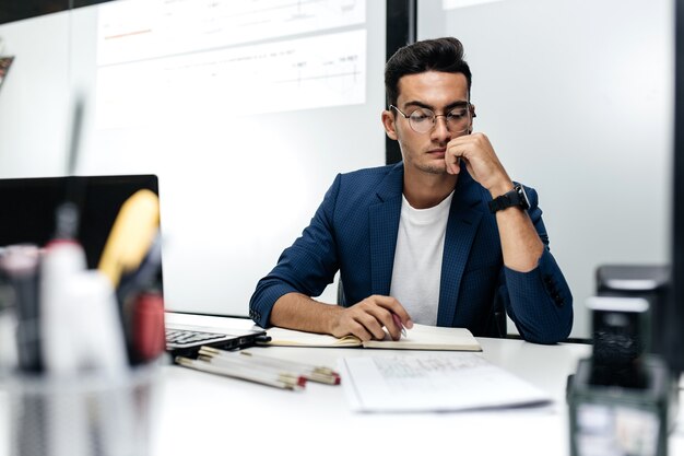 Joven arquitecto de pelo oscuro con gafas y una chaqueta azul está trabajando con documentos en el escritorio de la oficina.