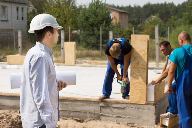 Foto joven arquitecto con un plano enrollado debajo del brazo de pie mirando a los trabajadores de la construcción en un sitio en construcción