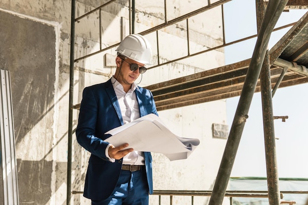 Joven arquitecto vistiendo traje formal y casco durante el control de la construcción de edificios sosteniendo un plano en un sitio de construcción