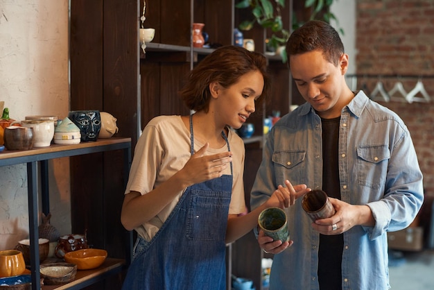 Joven artesana mostrando al cliente masculino dos tazas hechas a mano