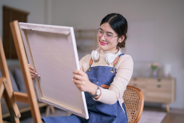 Una joven artista asiática lleva auriculares en el cuello y mira obras de arte en el lienzo con felicidad