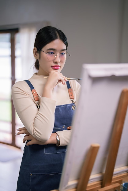 Una joven artista asiática sostiene un pincel y mira el lienzo para pensar y crear obras de arte de pintura