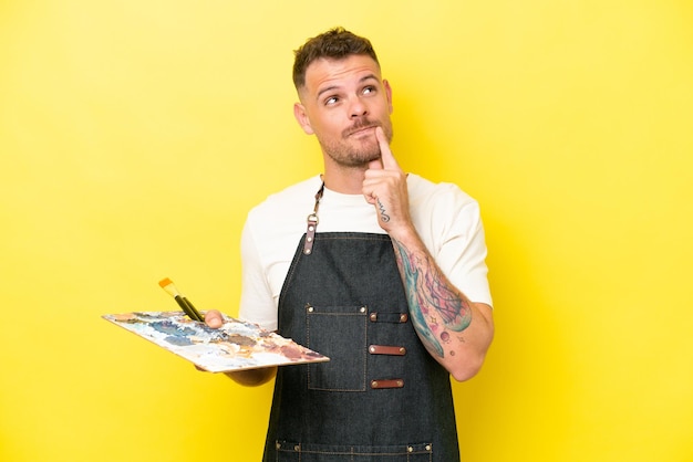 Joven artista caucásico sosteniendo una paleta aislada de fondo amarillo que tiene dudas mientras mira hacia arriba