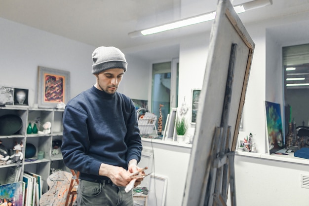 Joven artista está en un estudio cerca de un caballete con un lienzo con instrumentos en sus manos.