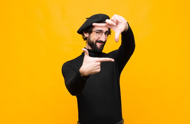 Joven artista francés hombre sintiéndose feliz, amable y positivo, sonriendo y haciendo un retrato o marco de fotos con las manos contra la pared naranja