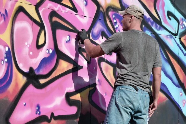 Joven artista de graffiti masculino caucásico dibujando una gran pintura de arte callejero en tonos azules y rosas