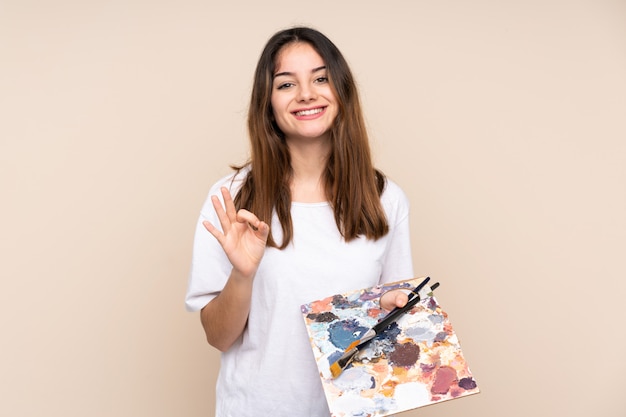 Joven artista hombre sosteniendo una paleta en color beige que muestra un signo bien con los dedos