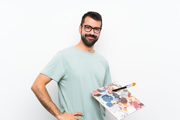 Joven artista hombre sosteniendo una paleta sobre pared aislada con gafas y sonriendo