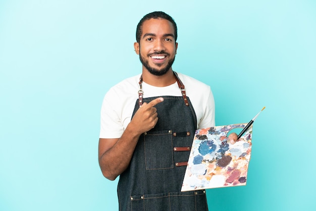 Joven artista latino sosteniendo una paleta aislada de fondo azul apuntando hacia un lado para presentar un producto