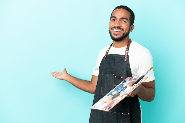 Joven artista latino sosteniendo una paleta aislada de fondo azul extendiendo las manos a un lado para invitar a venir