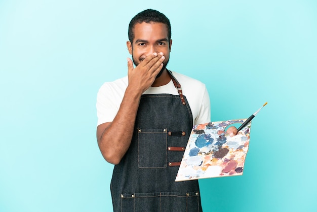 Joven artista latino sosteniendo una paleta aislada de fondo azul feliz y sonriente cubriendo la boca con la mano