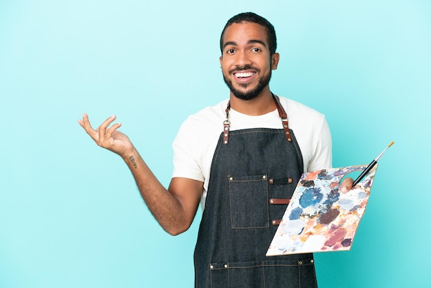 Joven artista latino sosteniendo una paleta aislada sobre fondo azul extendiendo las manos hacia el lado para invitar a venir