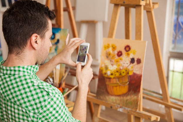 Joven artista masculino tomando una foto de su obra de arte con un teléfono inteligente para compartirla en una red social y venderla en línea