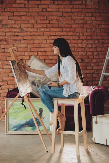 Una joven artista morena sonriente en su estudio está sosteniendo un pincel.