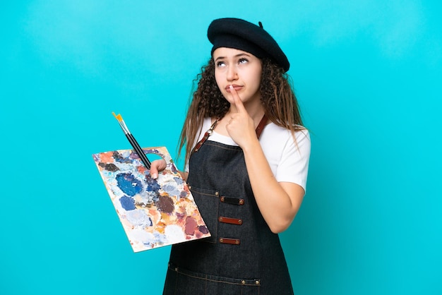 Joven artista mujer árabe sosteniendo una paleta aislada de fondo azul que tiene dudas mientras mira hacia arriba