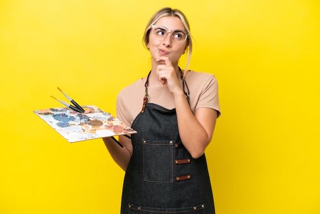 Joven artista mujer caucásica sosteniendo una paleta aislada de fondo amarillo y mirando hacia arriba