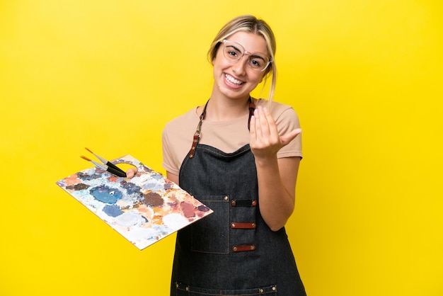 Joven artista mujer caucásica sosteniendo una paleta aislada sobre fondo amarillo invitando a venir con la mano Feliz de que hayas venido