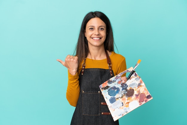 Joven artista mujer caucásica sosteniendo una paleta aislada sobre fondo azul apuntando hacia el lado para presentar un producto