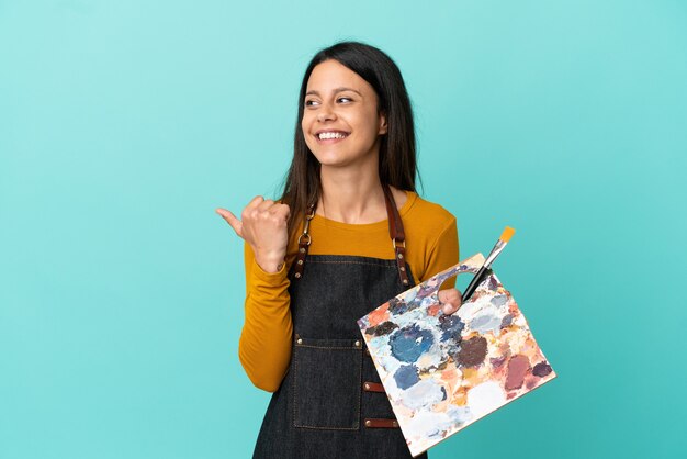 Joven artista mujer caucásica sosteniendo una paleta aislada sobre fondo azul apuntando hacia el lado para presentar un producto