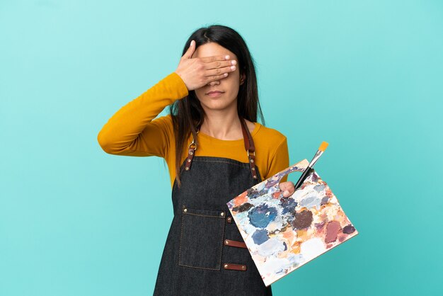 Joven artista mujer caucásica sosteniendo una paleta aislada sobre fondo azul cubriendo los ojos con las manos. No quiero ver algo