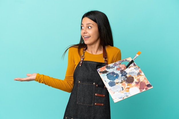 Joven artista mujer caucásica sosteniendo una paleta aislada sobre fondo azul con expresión de sorpresa mientras mira de lado